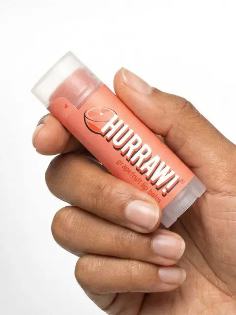 Hand holding a pink grapefruit lip balm tube with "Hurraw!" text.
