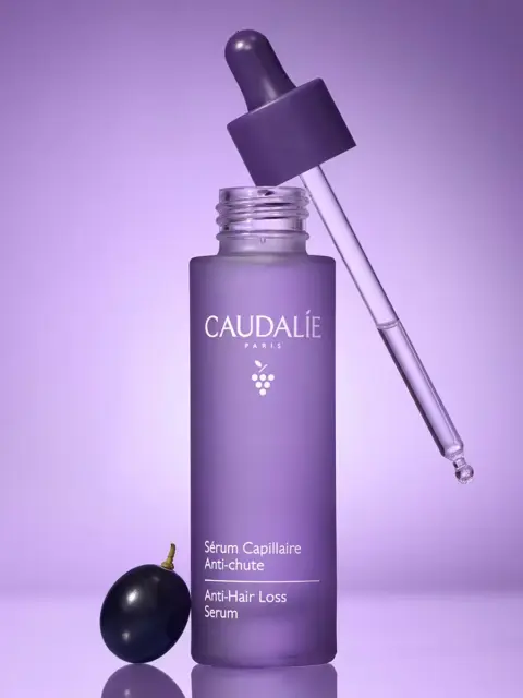 Purple Caudalie Anti-Hair Loss Serum bottle with dropper and grape on purple background.