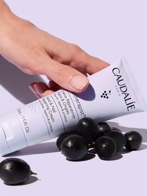 Hand holding Caudalie hand cream tube with black grapes on a light purple background.