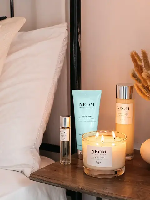 Neom products on bedside table with lit candle and dried flowers.