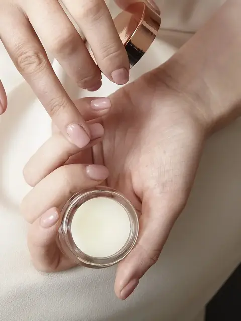 Hands holding a small jar of cream, with fingers applying it.