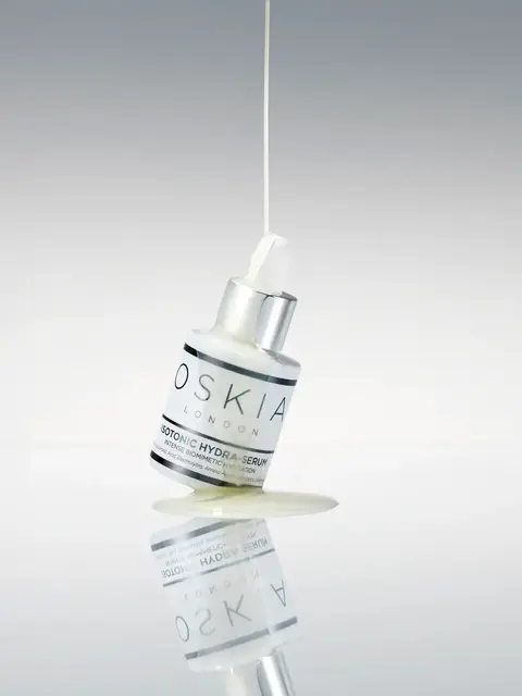 Oskia Isotonic Hydra-Serum bottle with liquid pouring, reflected on a glossy surface.
