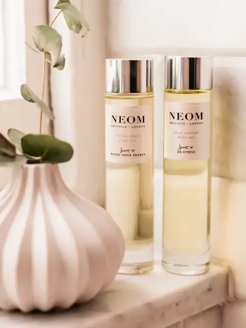 Two Neom Organics body oils on a marble shelf beside a white vase with green leaves.