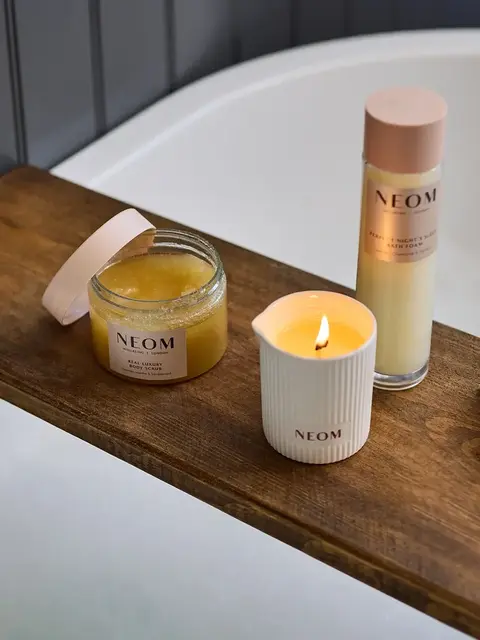 Neom body scrub, bath foam, and lit candle on wooden bath tray.
