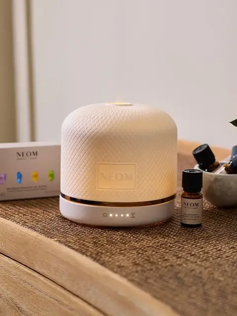 White NEOM diffuser on a wooden table with essential oils and packaging.