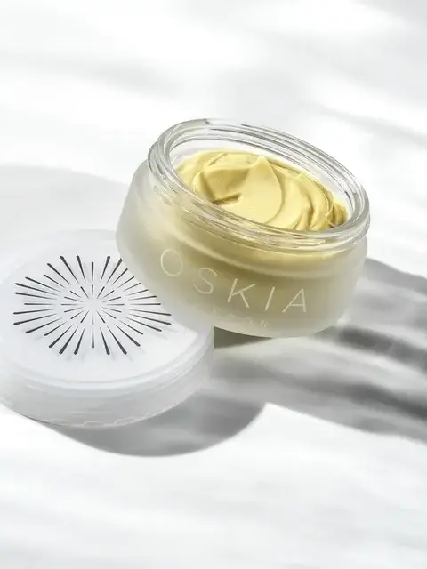 Open jar of yellow cream with "OSKIA" on the side, lid beside it on a white surface.
