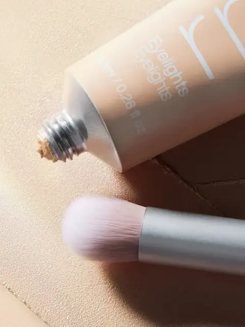 Open tube of Eyelights cream with a makeup brush on a textured surface.