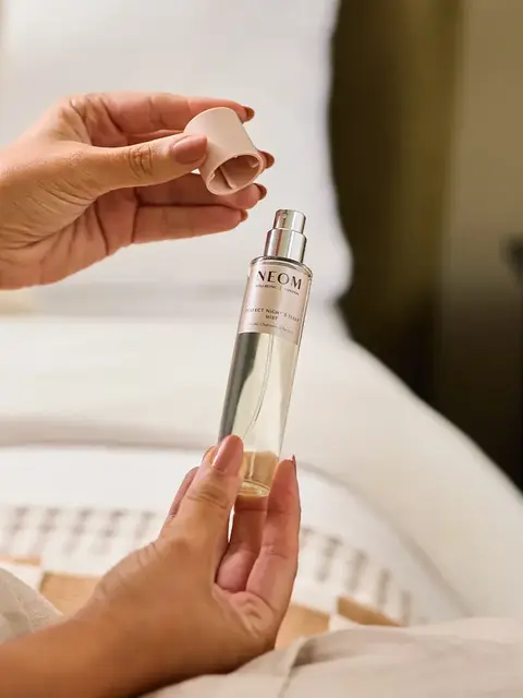 Hands holding a NEOM sleep spray bottle with cap removed, near a bed.