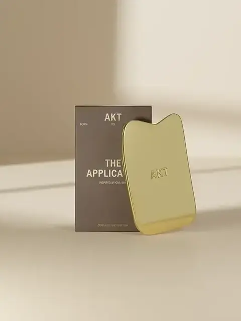 Gold AKT applicator with packaging on a beige surface.