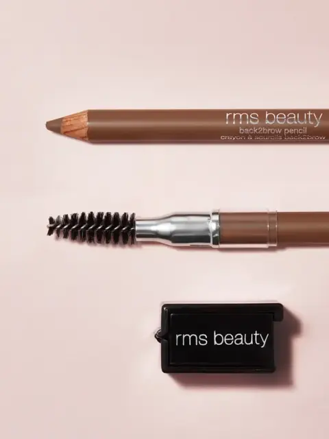 Brown eyebrow pencil, spoolie brush, and sharpener on a light pink background. Text: "rms beauty".