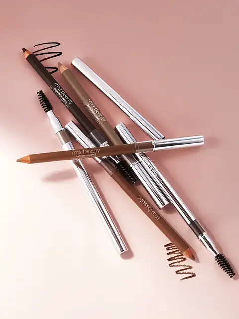 Assorted RMS Beauty brow pencils on a pink surface.