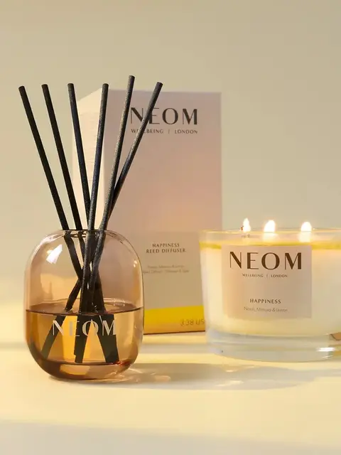 NEOM Happiness reed diffuser and lit candle on a light surface.