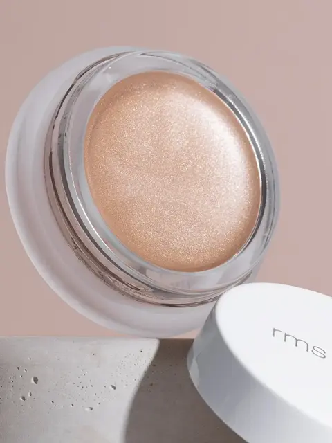 Open jar of shimmering peach cream with "rms beauty" on the white lid, set on a stone surface.