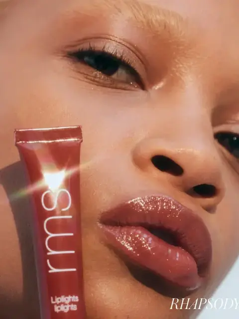 Person with glossy lips holding RMS Liplights tube. Text: "RHAPSODY".
