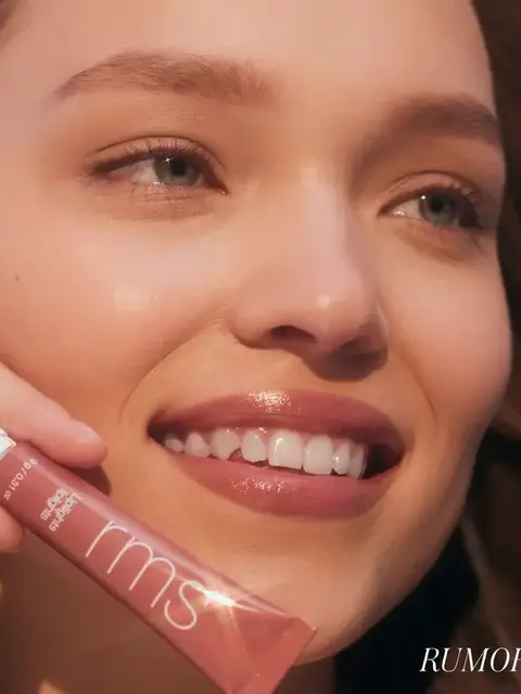 Smiling person holding RMS Beauty lip gloss tube. Text: "RUMOR".