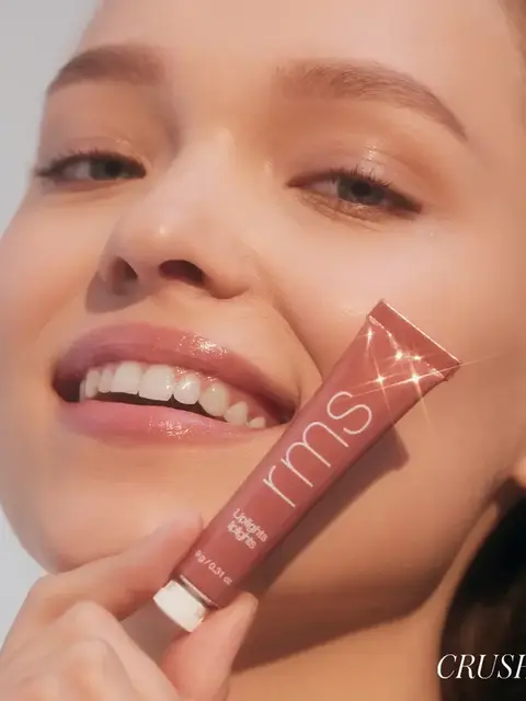 Smiling person holding a tube of RMS Beauty lip gloss labelled "Crush".
