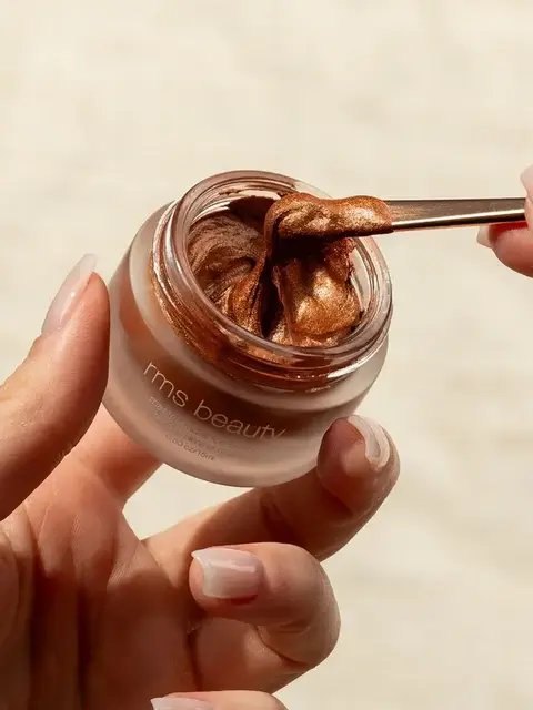Hands holding a jar of rms beauty bronze cream with a spatula.