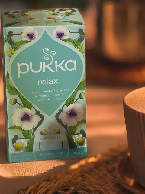 Pukka Relax herbal tea box with chamomile, fennel, and marshmallow root, next to a cup on a woven mat.