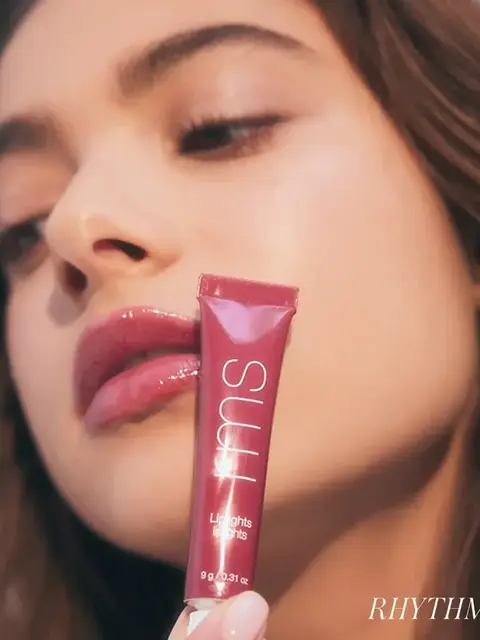 Woman holding a pink lip gloss tube labelled "SUU" near her face.