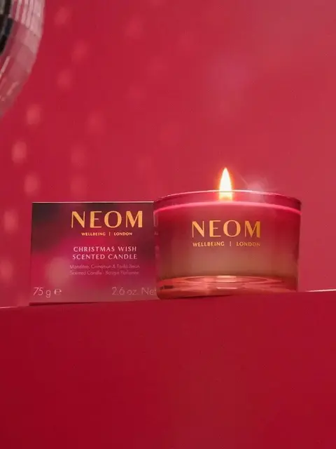 NEOM Christmas Wish scented candle lit, with packaging, on a red background.