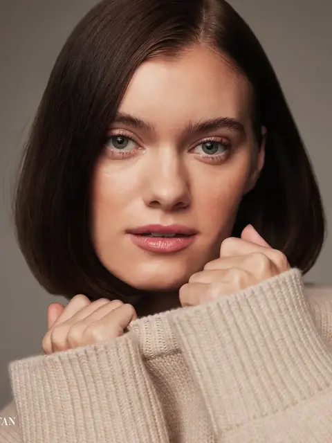 Woman with short brown hair in a beige jumper, holding collar near face. Text: "TAN".