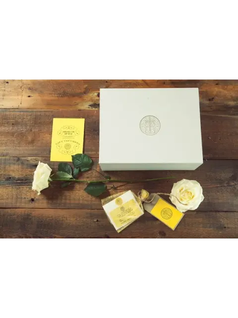 White gift box with logo, yellow card, soap, and white roses on wooden table.