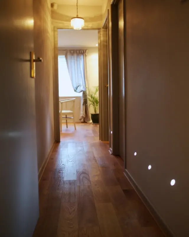 Hallway with wooden floor, open door, and a lit room with a chair and plant visible at the end.