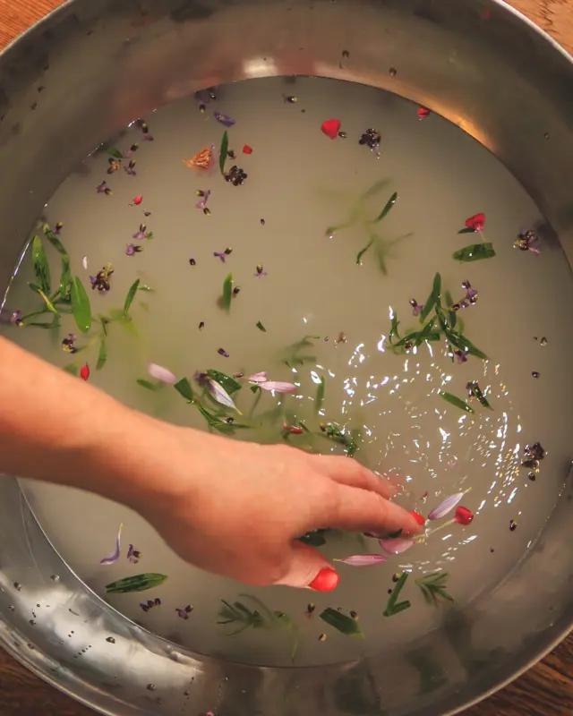 Hand stirring water with petals and herbs in a metal bowl on a wooden surface.