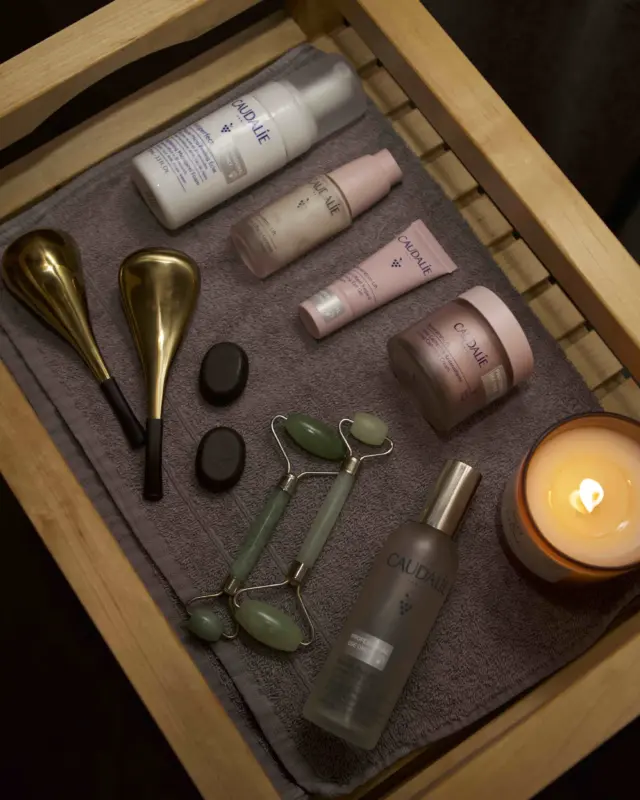 Skincare products, jade roller, candle, and gold spoons on a wooden tray with a grey towel.