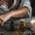 Person applying oil to arm with dropper, surrounded by bottles, candle, and flowers on wooden table.
