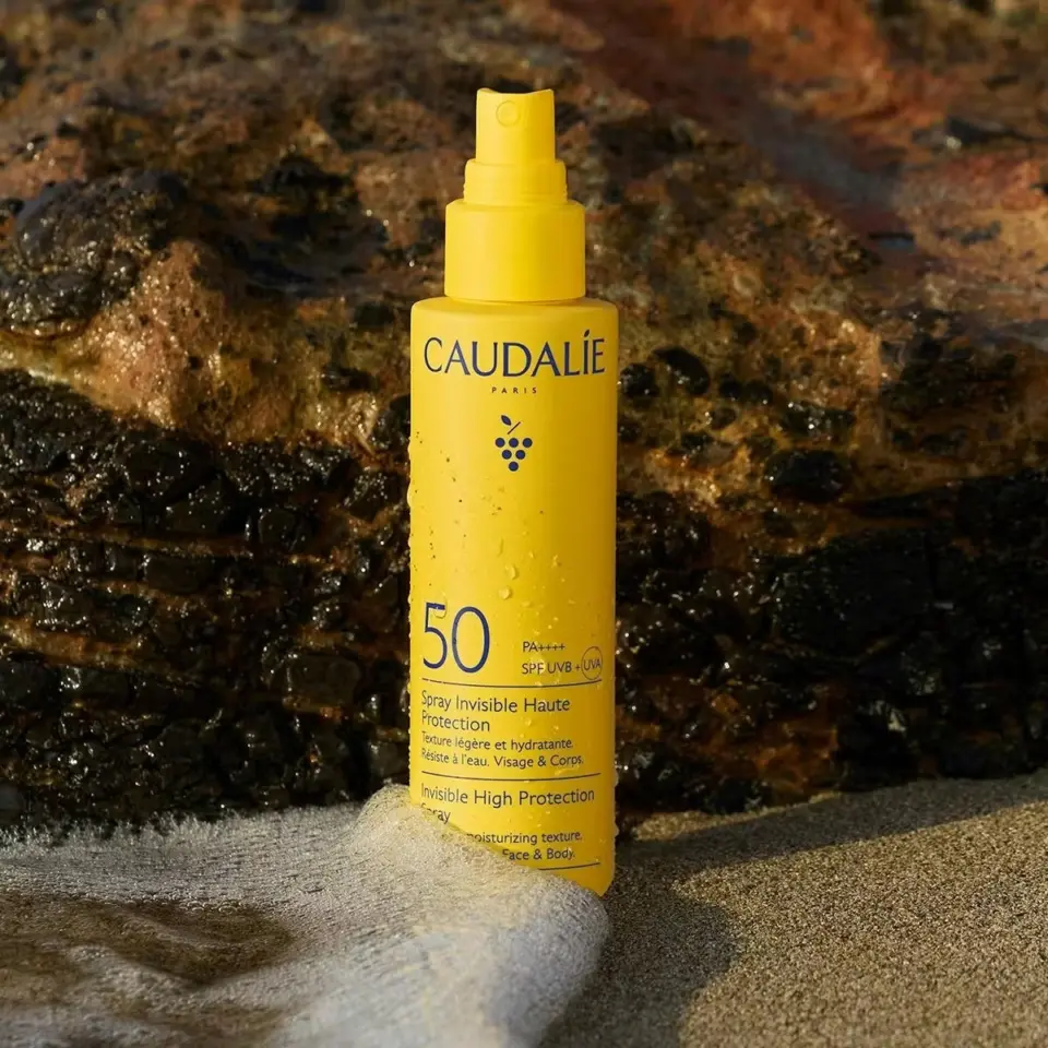Yellow Caudalie SPF 50 spray bottle on a sandy beach with rocks in the background.