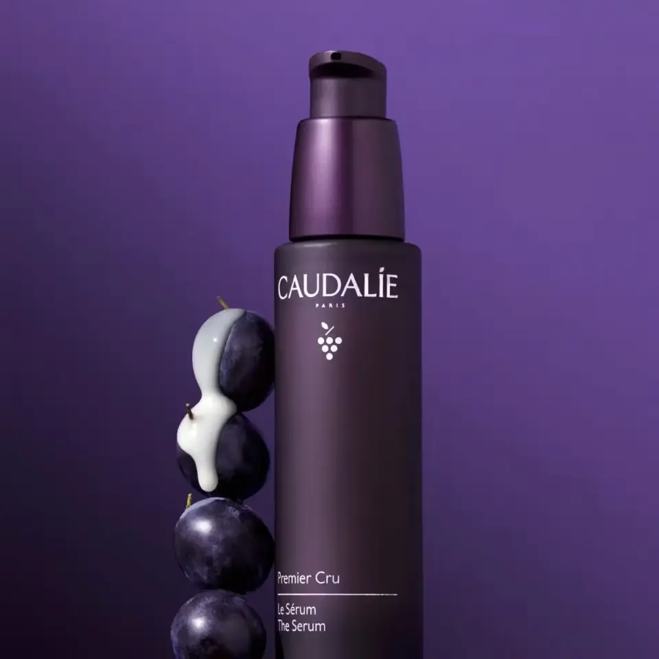 Caudalie Premier Cru Serum bottle with grapes and cream on purple background.
