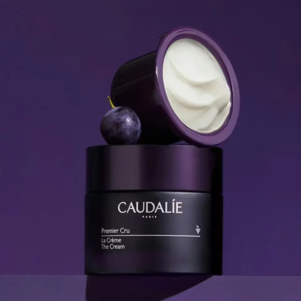 Caudalie Premier Cru cream jar with lid open, next to a grape, on a purple background.