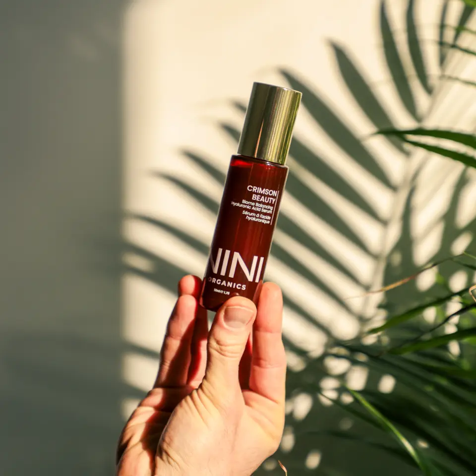 Hand holding NINI Organics Crimson Beauty serum, with plant shadow in background.