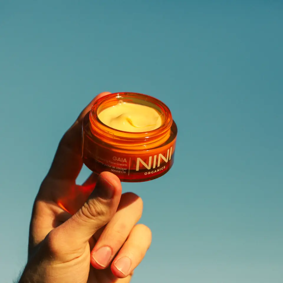 Hand holding an open jar of NINI Organics cream against a clear blue sky.