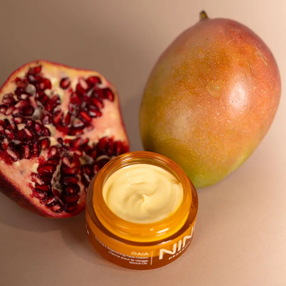 Open jar of cream with pomegranate half and whole mango on beige background.