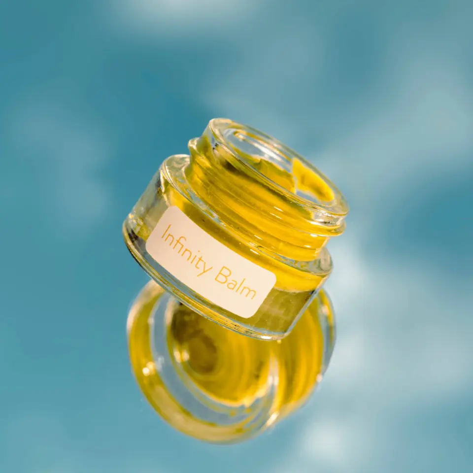 Glass jar of Infinity Balm with yellow contents against a blue background.