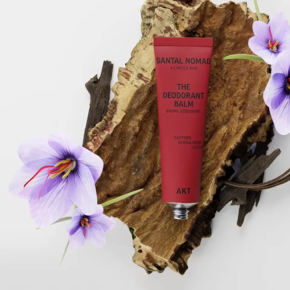 Red deodorant balm tube on bark with purple flowers. Text: Santal Nomad, AKT, saffron, sandalwood, leather.
