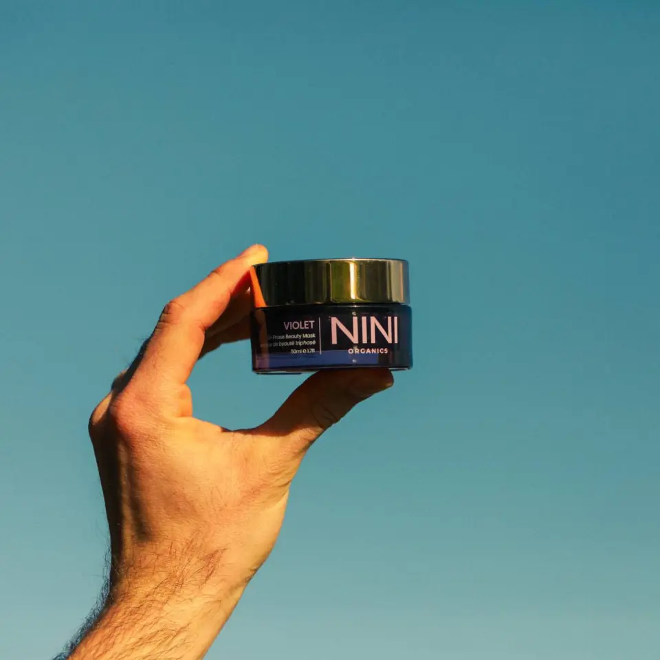 Hand holding NINI Organics Violet Cream against a clear blue sky.