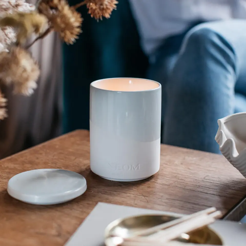 Lit white candle in a ceramic holder on a wooden table, with a lid beside it.