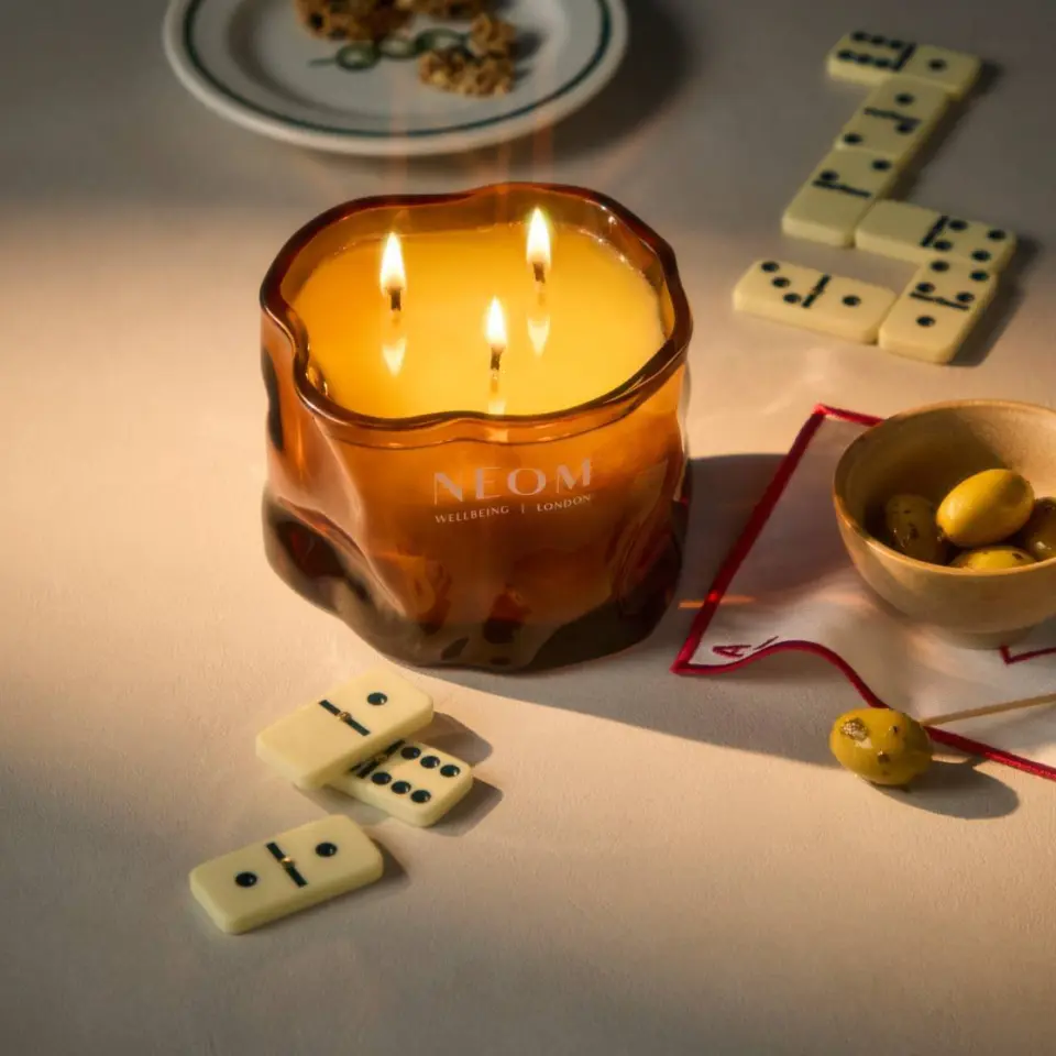Lit NEOM candle with dominoes, olives in a bowl, and a plate of snacks on a table.