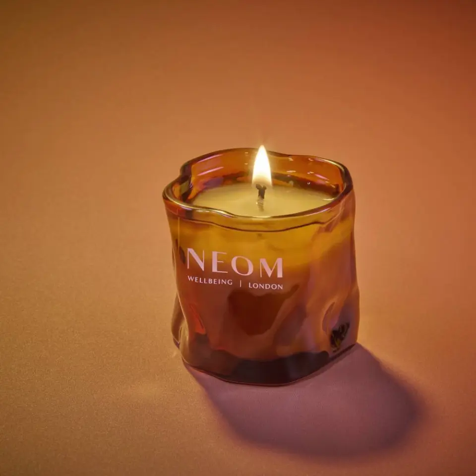 Lit candle in amber glass holder with "NEOM Wellbeing London" text. Warm background.