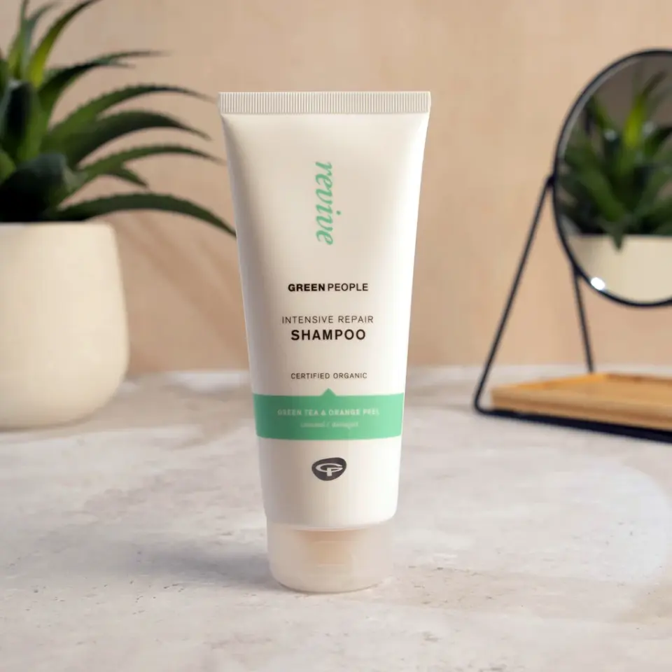 Green People Intensive Repair Shampoo tube on a bathroom counter with plants and a mirror.