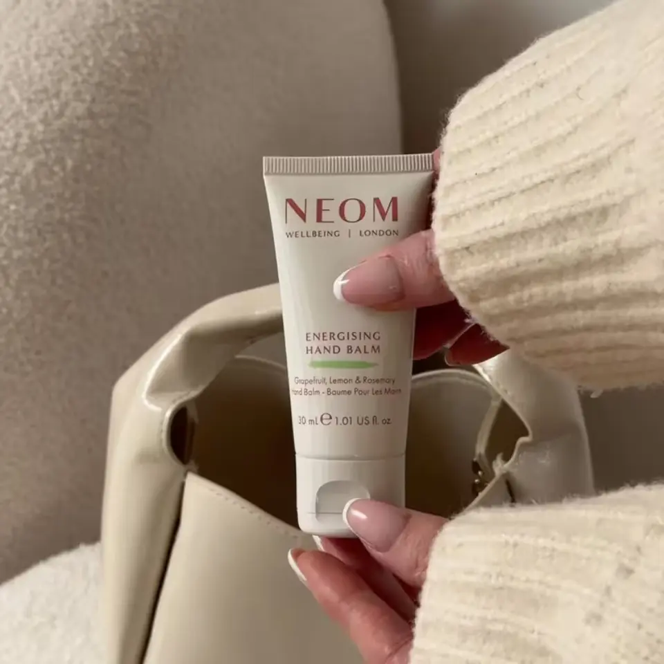 NEOM Energising Hand Balm, 30ml, held by a person in a cream jumper, near a beige bag.