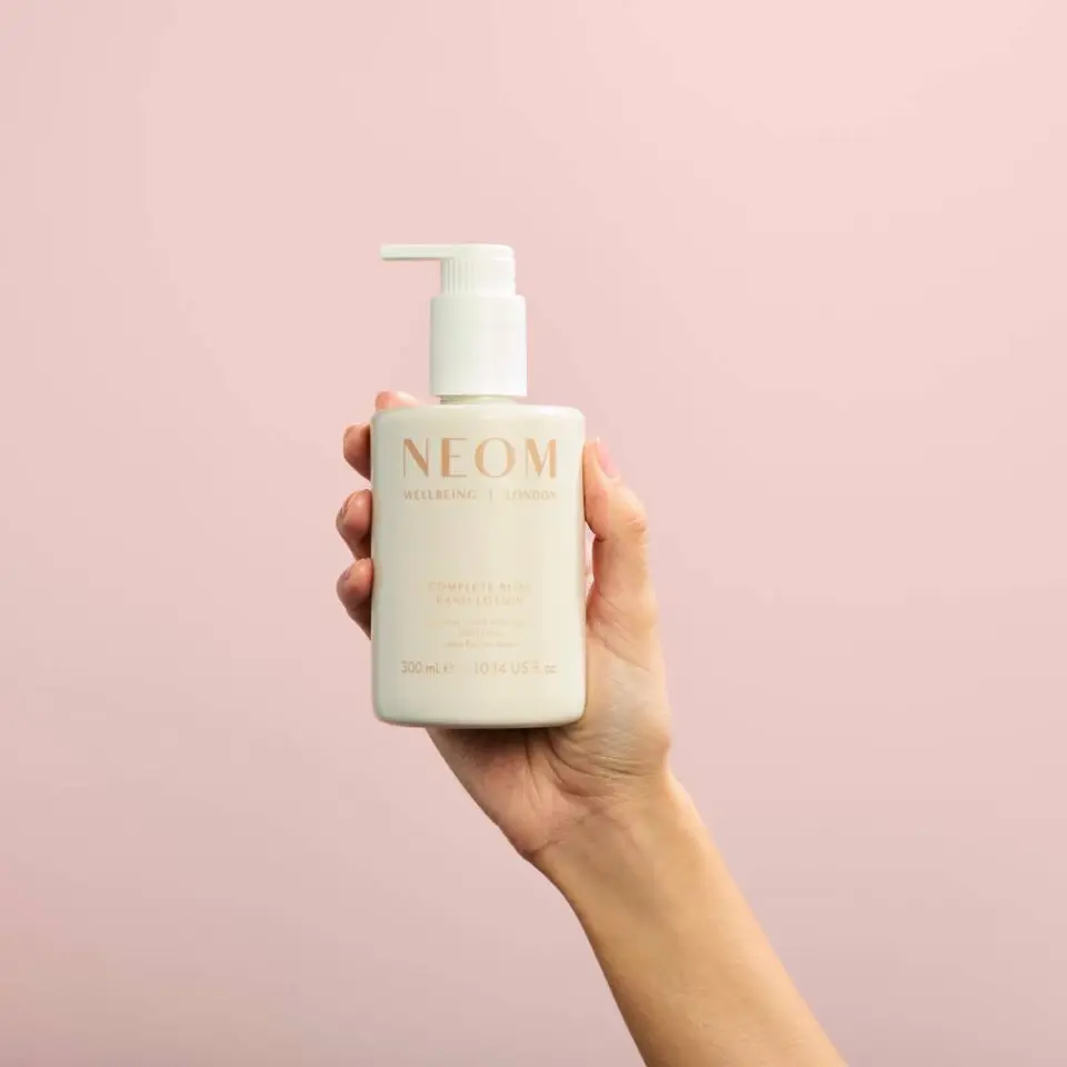 Hand holding NEOM lotion bottle against a pink background.