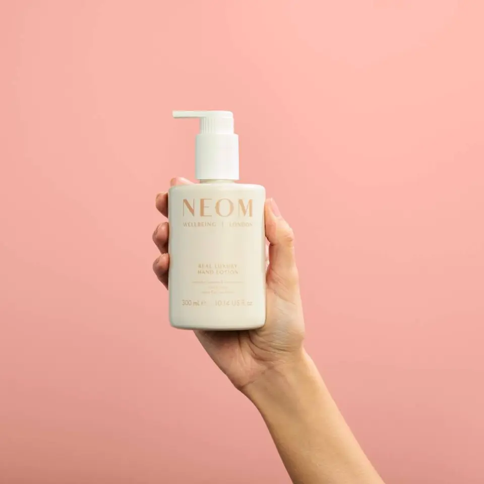 Hand holding NEOM hand lotion bottle against pink background.