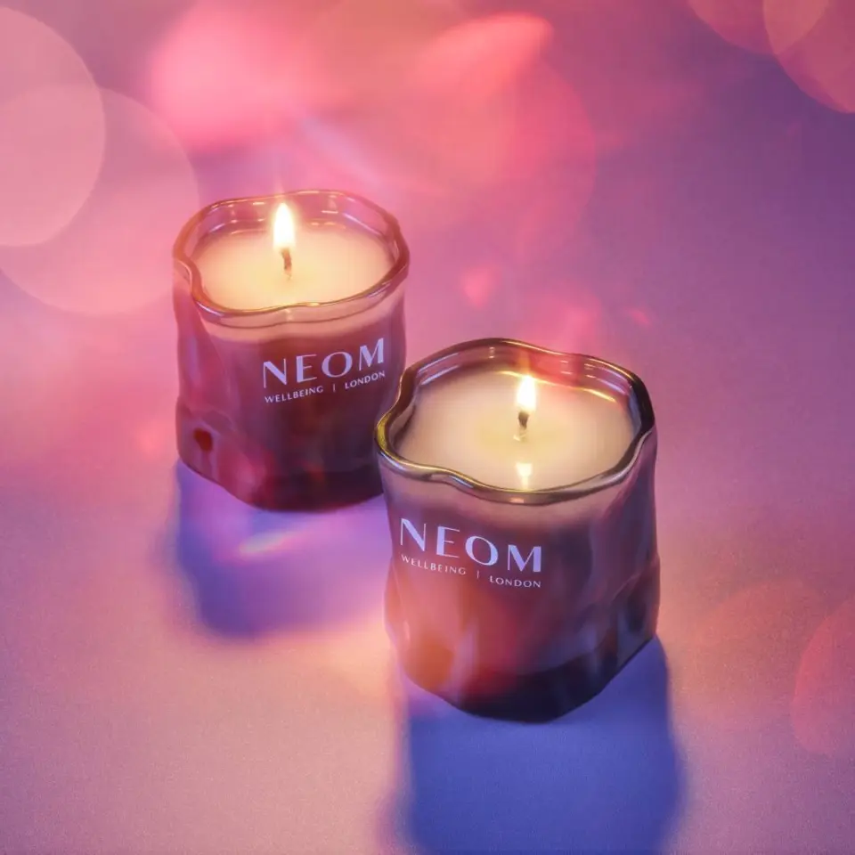 Two lit NEOM candles on a purple surface with soft pink bokeh lights.