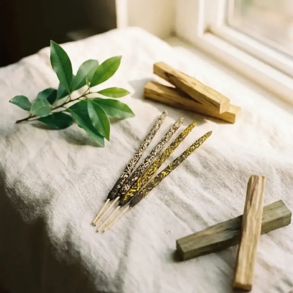 Incense sticks, wooden blocks, and green leaves on a linen cloth by a window.
