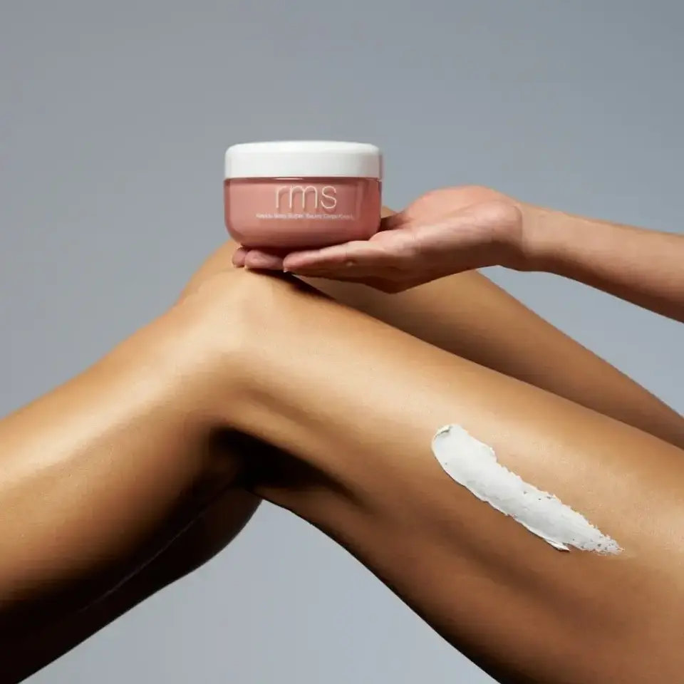 Hand holding RMS body butter above a leg with cream applied.