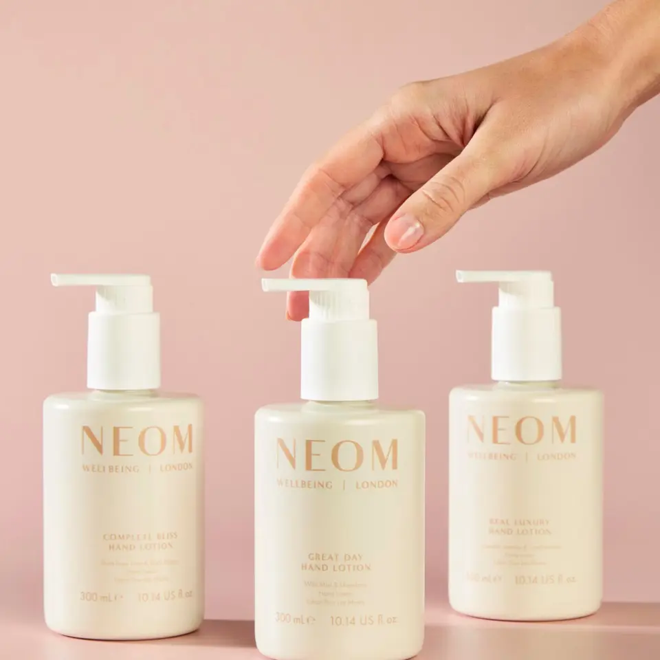 Three NEOM hand lotions on pink background, hand reaching for centre bottle.
